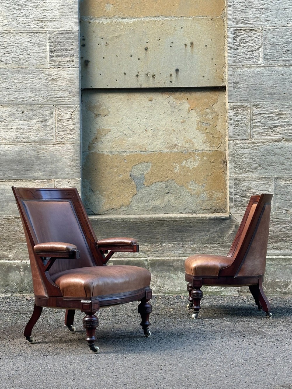 A pair of nineteenth-century Greek Revival mahogany armchairs upholstered in warm brown leather with turned front legs on castors and incised geometric detailing.