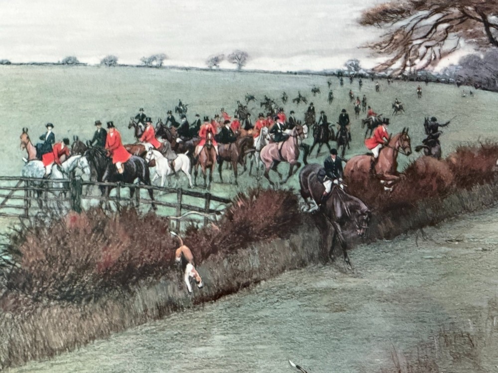 A panoramic antique colour print depicting a fox hunt in full pursuit across a green countryside landscape with riders jumping a hedge and hounds running in the foreground. View II
