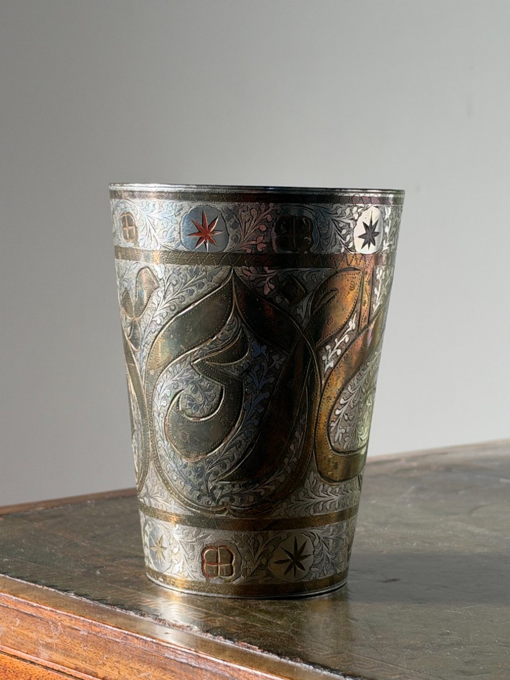 A tapered white metal beaker featuring detailed hand-engraved foliage motifs, star emblems, and a tarnished patina.