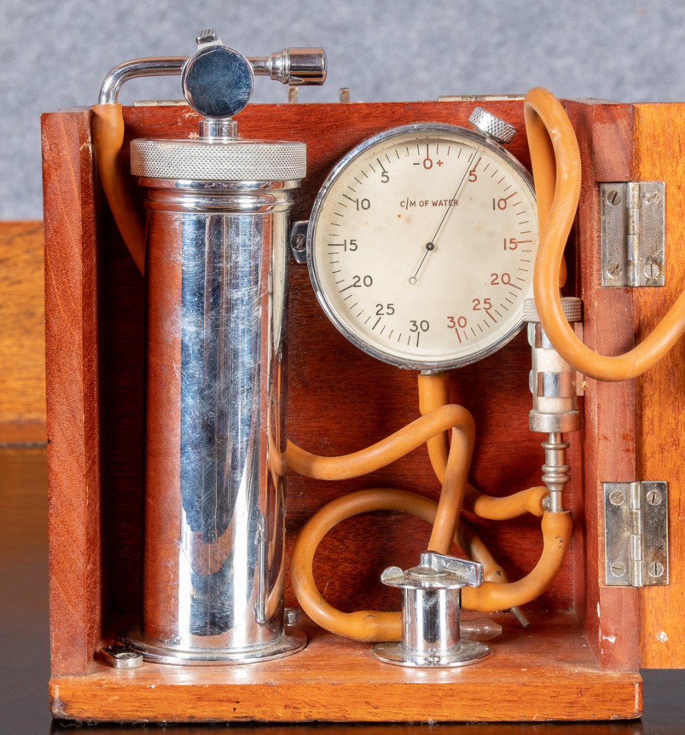 A vintage pneumothorax medical device housed inside an open fitted wooden box, featuring a chrome cylinder, flexible orange rubber tubing, and a round dial gauge marked in centimetres of water.