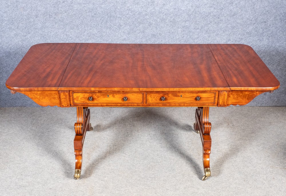 A Regency mahogany sofa table with two frieze drawers, drop-leaf ends, curved trestle supports, and brass castors.