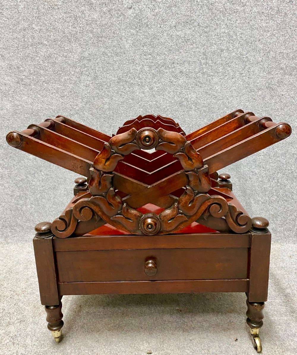 An antique mahogany music canterbury featuring carved acanthus side rails, slatted dividers, a single lower drawer with a central knob handle, and turned legs set on brass castors.