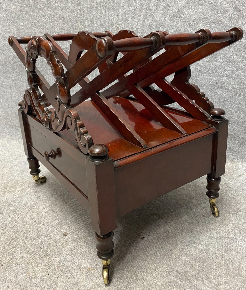 An antique mahogany music canterbury featuring carved acanthus side rails, slatted dividers, a single lower drawer with a central knob handle, and turned legs set on brass castors. View II