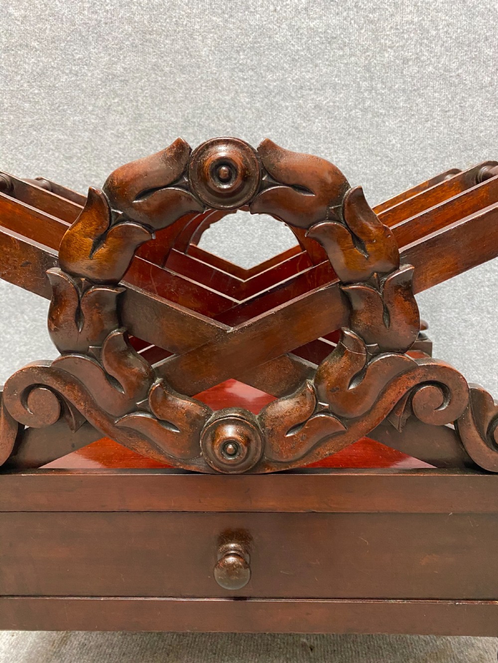 An antique mahogany music canterbury featuring carved acanthus side rails, slatted dividers, a single lower drawer with a central knob handle, and turned legs set on brass castors. View III