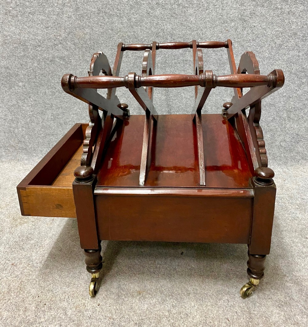 An antique mahogany music canterbury featuring carved acanthus side rails, slatted dividers, a single lower drawer with a central knob handle, and turned legs set on brass castors. View IV