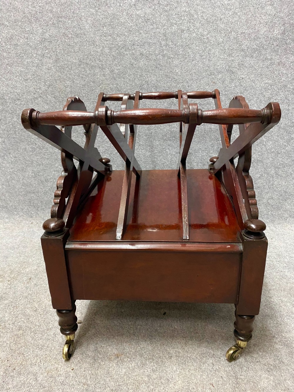 An antique mahogany music canterbury featuring carved acanthus side rails, slatted dividers, a single lower drawer with a central knob handle, and turned legs set on brass castors. View V