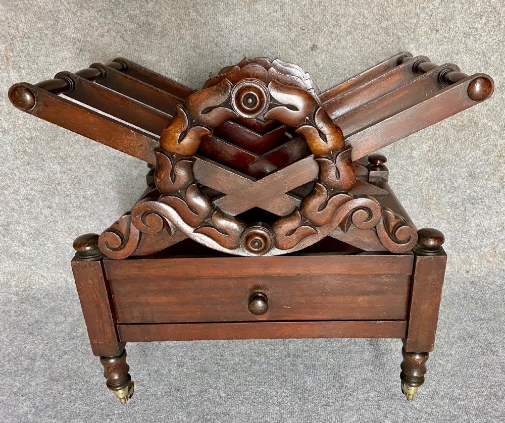 An antique mahogany music canterbury featuring carved acanthus side rails, slatted dividers, a single lower drawer with a central knob handle, and turned legs set on brass castors. View VII