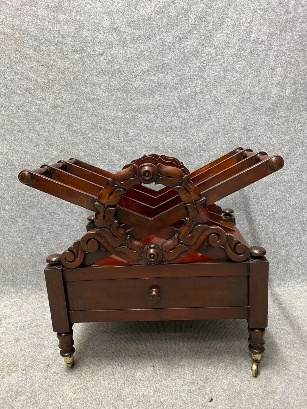 An antique mahogany music canterbury featuring carved acanthus side rails, slatted dividers, a single lower drawer with a central knob handle, and turned legs set on brass castors. View IX