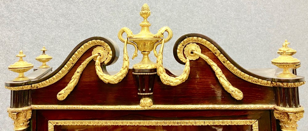 A two-tier dark mahogany display cabinet featuring gilt bronze mounts, glazed panels, fluted columns, and finial decorations. View VI