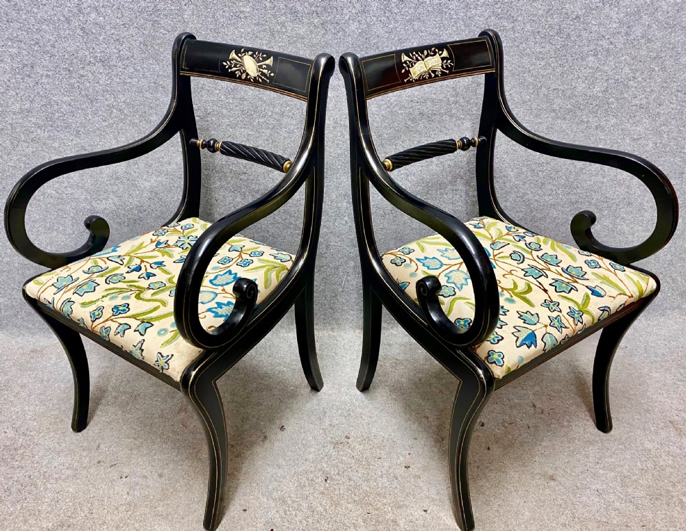 A pair of black ebonised Regency style armchairs featuring gold gilt classical motifs on the top rails, swept scrolled arms, and botanical fabric seats.