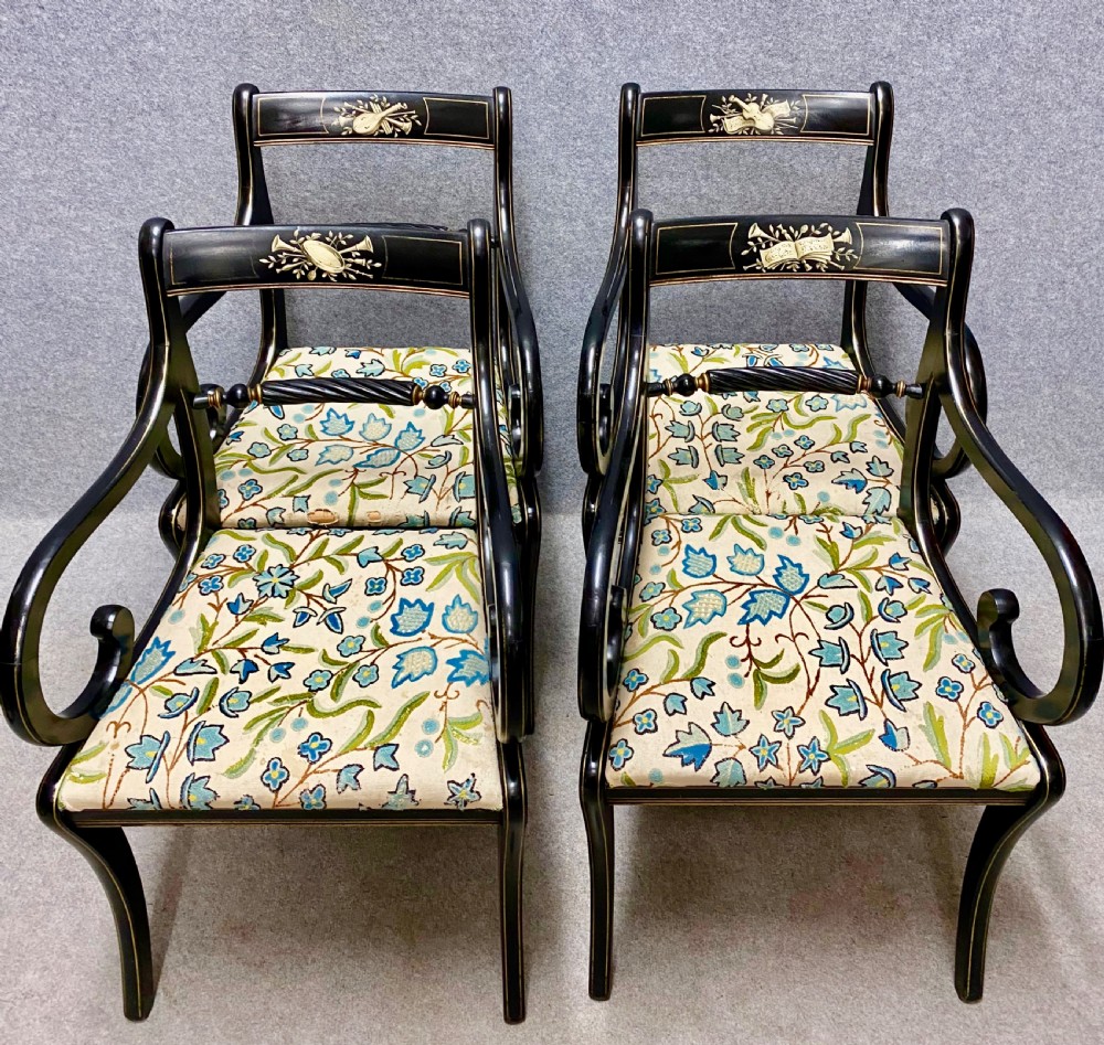 A set of four ebonised Regency style elbow chairs featuring sweeping armrests, hand-painted classical crest rails, and floral upholstered seats.