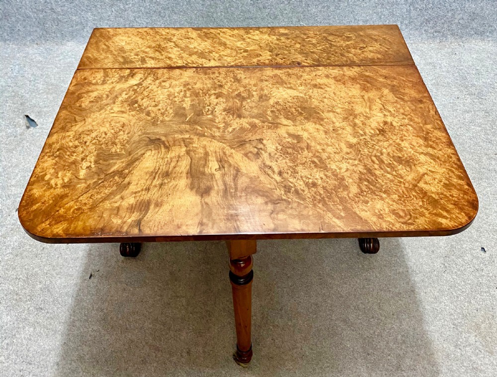 A Victorian burr walnut Sutherland table shown with its drop leaves closed, displaying swirling wood grain veneers, carved legs, and brass castors. View II