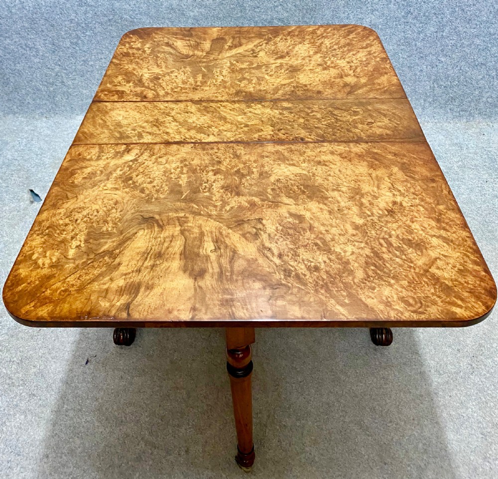 A Victorian burr walnut Sutherland table shown with its drop leaves closed, displaying swirling wood grain veneers, carved legs, and brass castors. View III