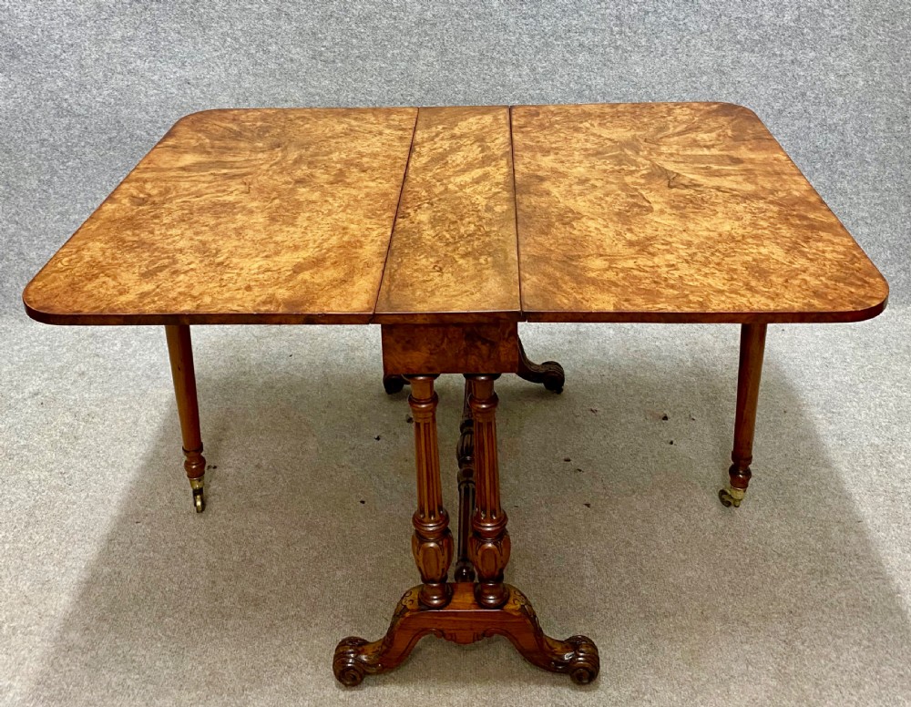 A Victorian burr walnut Sutherland table shown with its drop leaves closed, displaying swirling wood grain veneers, carved legs, and brass castors. View IV