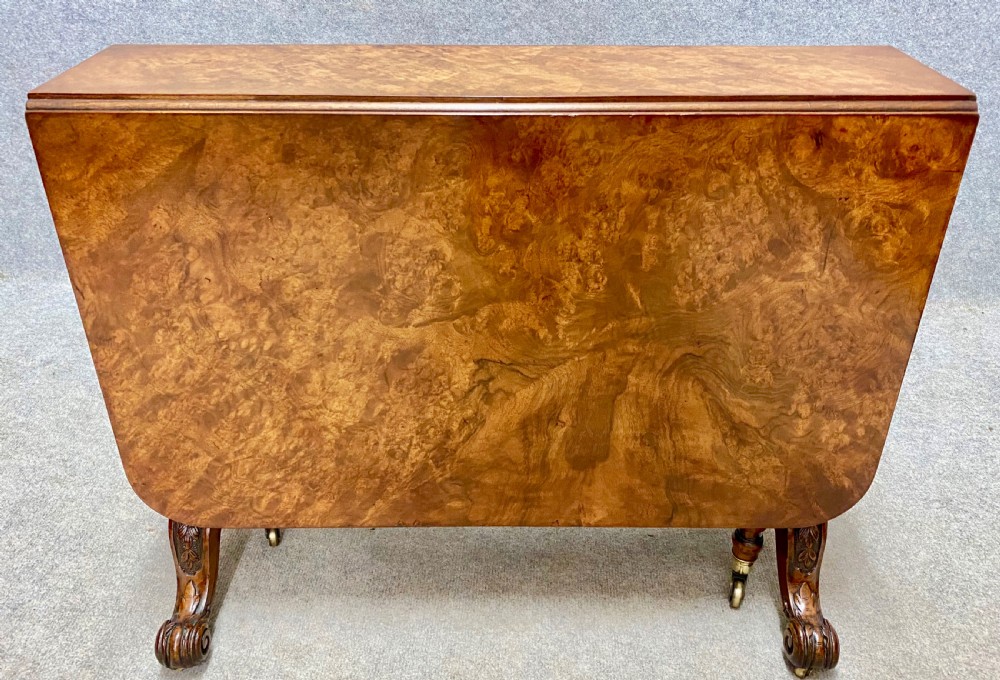 A Victorian burr walnut Sutherland table shown with its drop leaves closed, displaying swirling wood grain veneers, carved legs, and brass castors. View IX