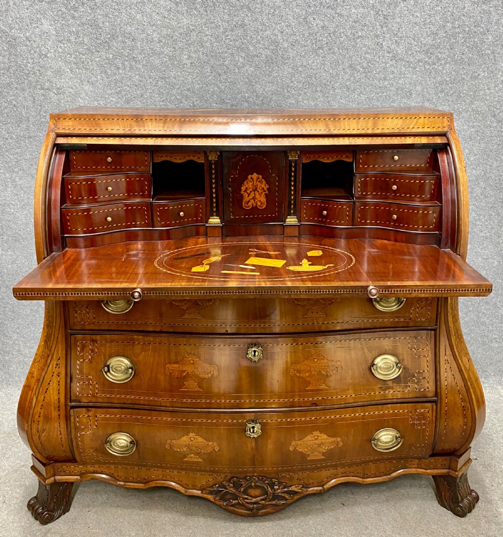 A Dutch marquetry cylinder bureau crafted from warm brown wood with trompe loeil writing tool inlays, fitted drawers, brass handles, and a curved bombe base.
