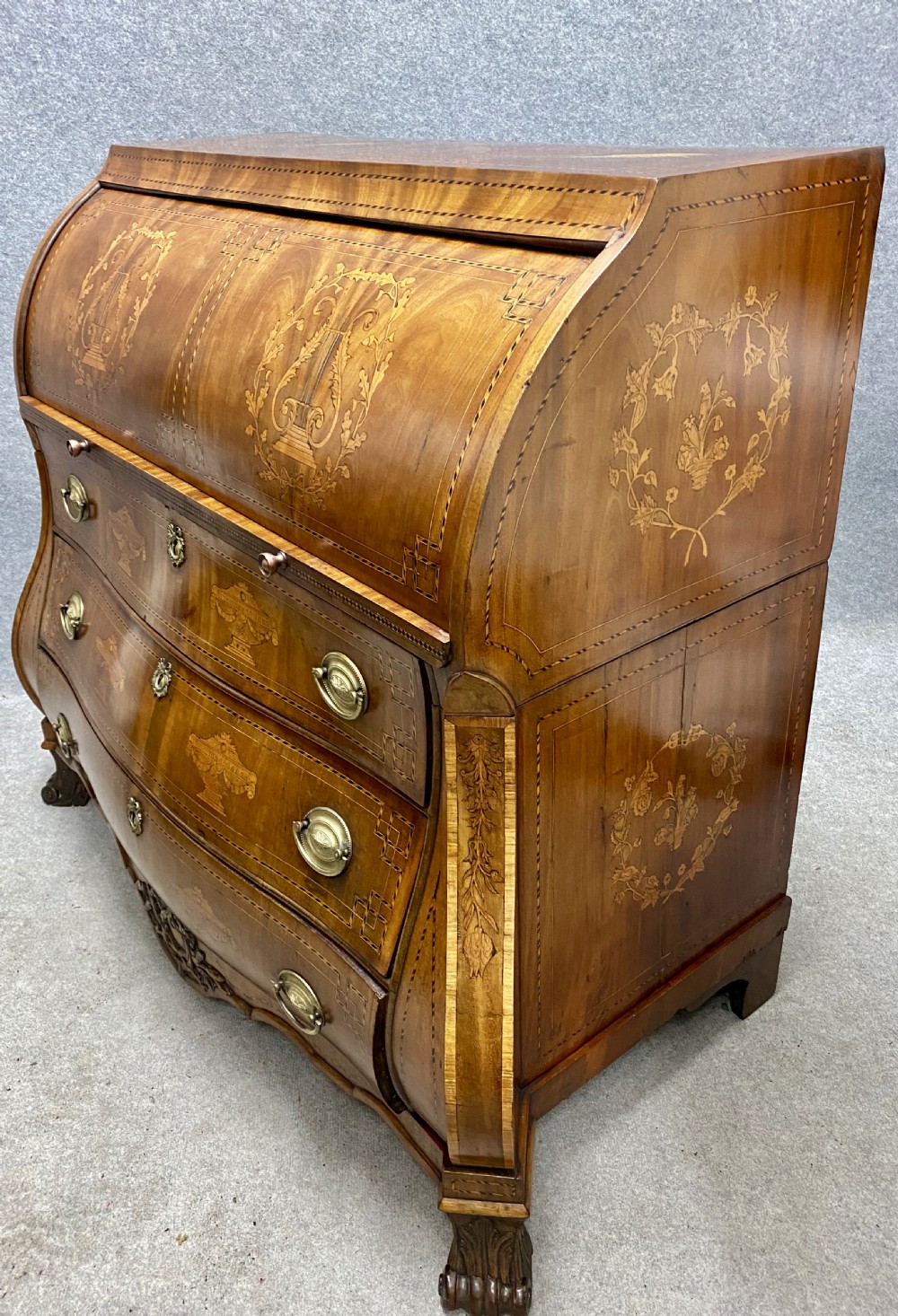 A Dutch marquetry cylinder bureau crafted from warm brown wood with trompe loeil writing tool inlays, fitted drawers, brass handles, and a curved bombe base. View II