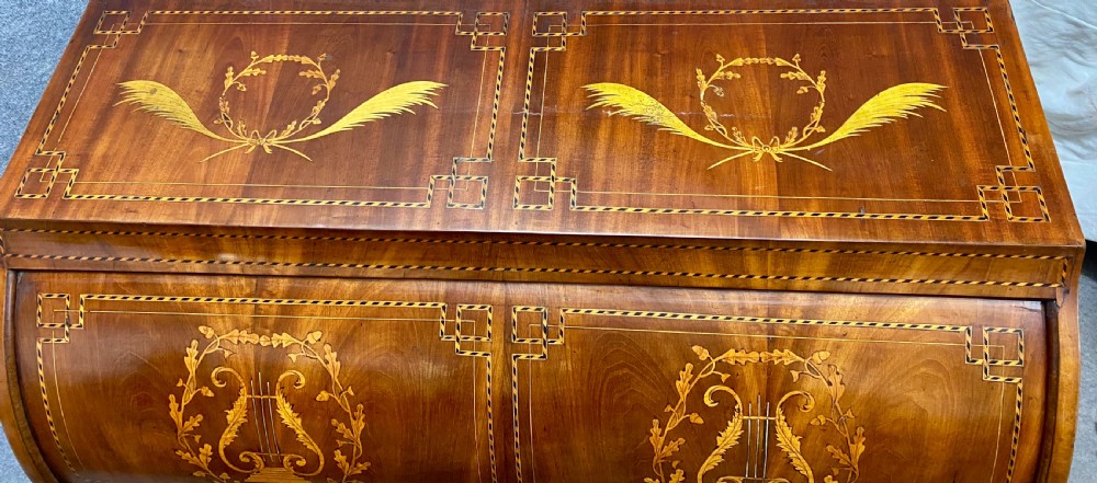 A Dutch marquetry cylinder bureau crafted from warm brown wood with trompe loeil writing tool inlays, fitted drawers, brass handles, and a curved bombe base. View VI