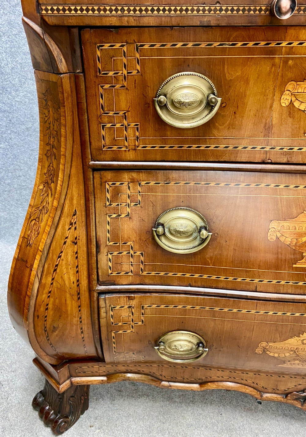 A Dutch marquetry cylinder bureau crafted from warm brown wood with trompe loeil writing tool inlays, fitted drawers, brass handles, and a curved bombe base. View VII
