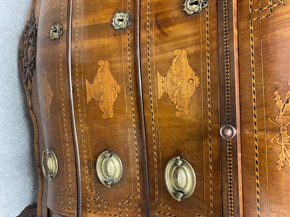 A Dutch marquetry cylinder bureau crafted from warm brown wood with trompe loeil writing tool inlays, fitted drawers, brass handles, and a curved bombe base. View X