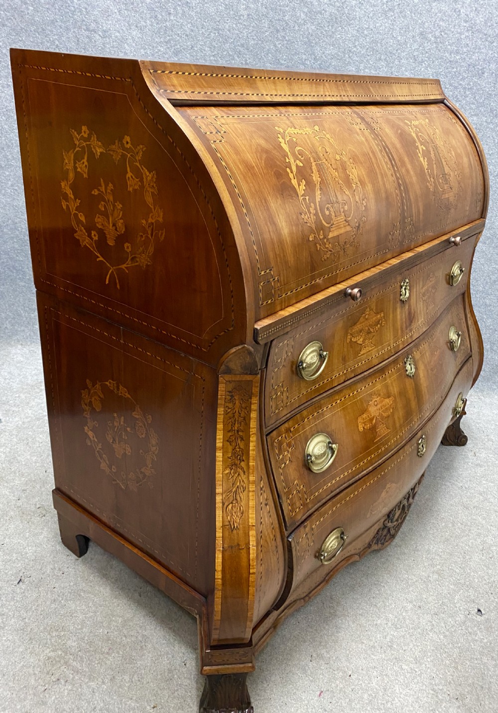 A Dutch marquetry cylinder bureau crafted from warm brown wood with trompe loeil writing tool inlays, fitted drawers, brass handles, and a curved bombe base. View XVIII