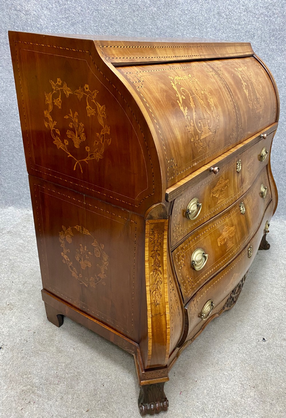 A Dutch marquetry cylinder bureau crafted from warm brown wood with trompe loeil writing tool inlays, fitted drawers, brass handles, and a curved bombe base. View XIX
