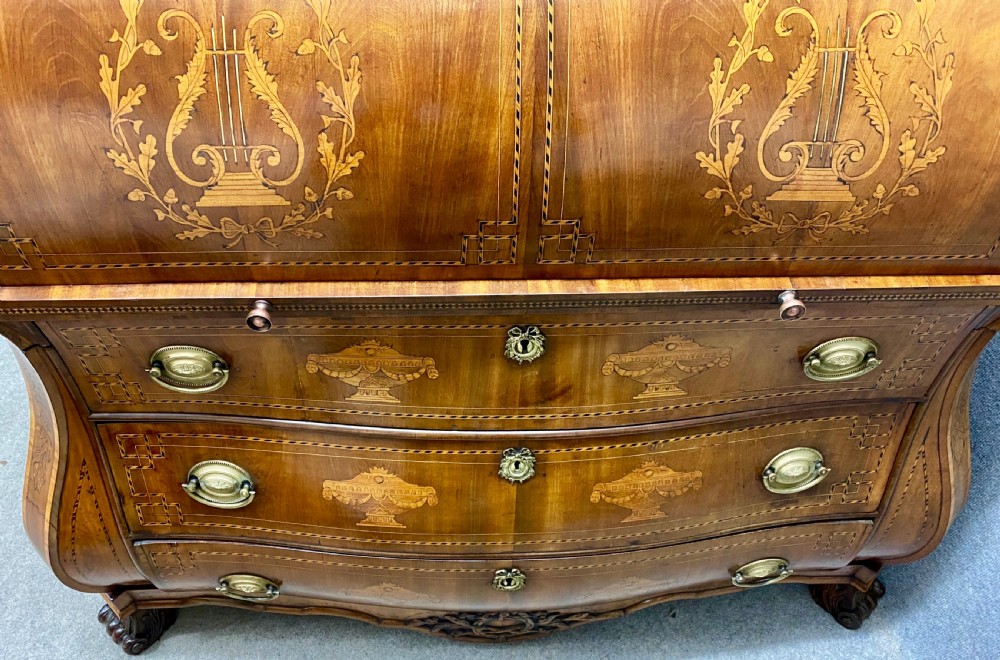 A Dutch marquetry cylinder bureau crafted from warm brown wood with trompe loeil writing tool inlays, fitted drawers, brass handles, and a curved bombe base. View XX
