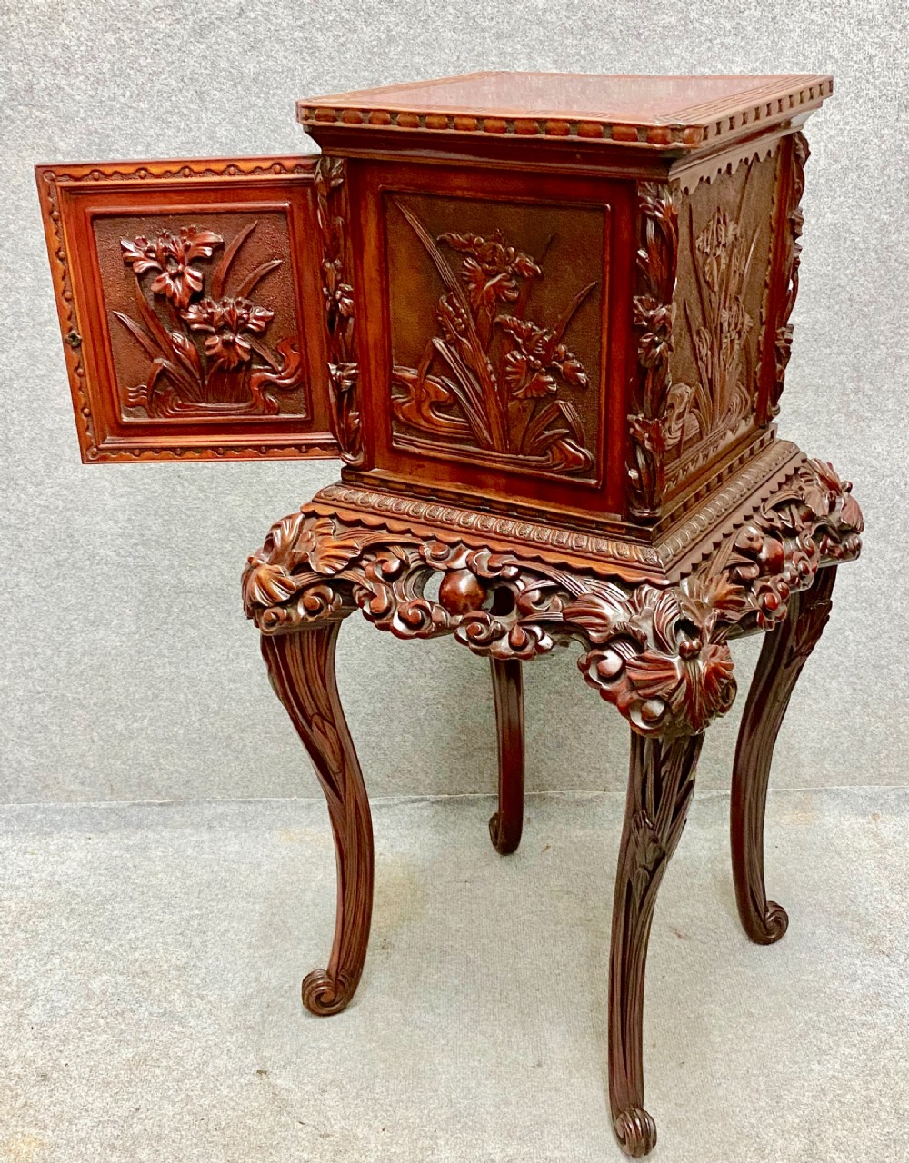 A reddish brown carved cinnabar lacquer cabinet with floral relief panels resting on a tall cabriole leg stand with an open side door.