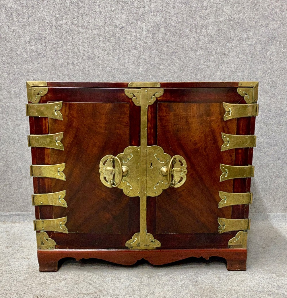 A small Chinese hardwood cabinet featuring polished dark wood, twin front doors, and decorative engraved brass mounts, hinges, and a central circular lockplate.