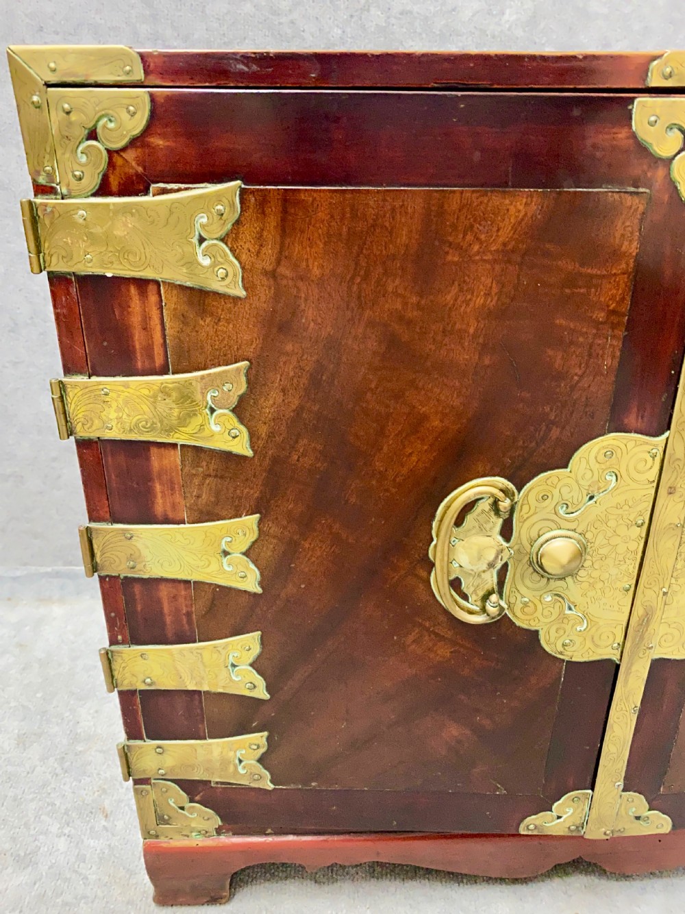 A small Chinese hardwood cabinet featuring polished dark wood, twin front doors, and decorative engraved brass mounts, hinges, and a central circular lockplate. View IV