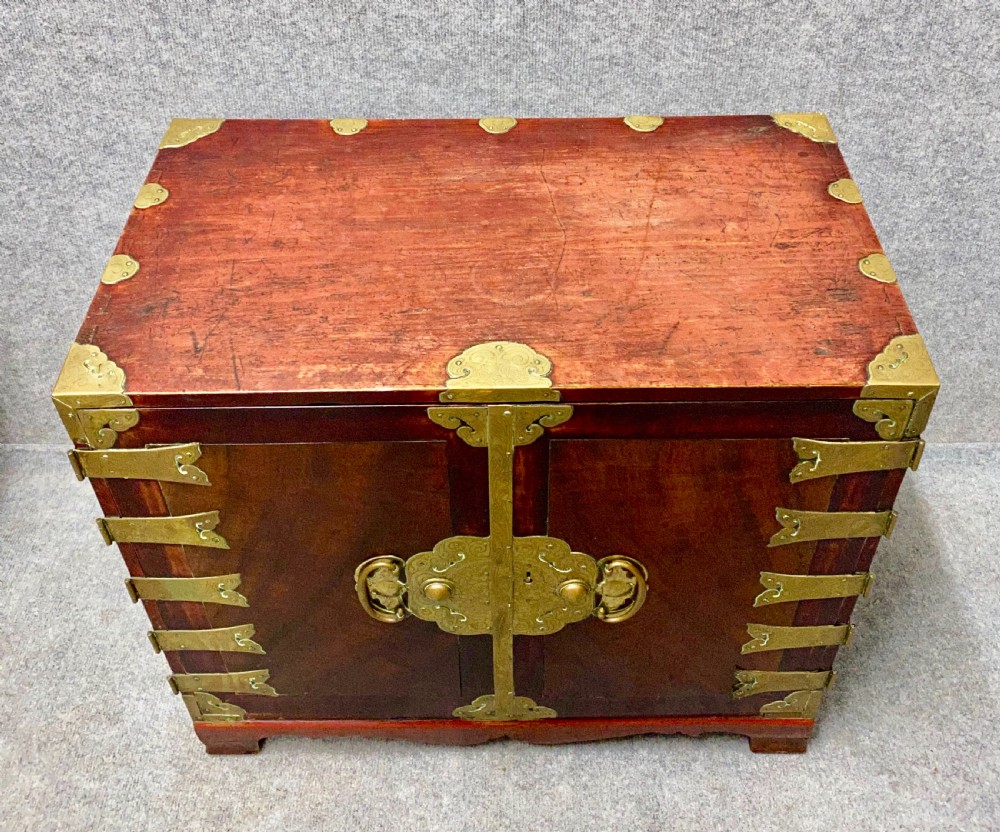 A small Chinese hardwood cabinet featuring polished dark wood, twin front doors, and decorative engraved brass mounts, hinges, and a central circular lockplate. View V