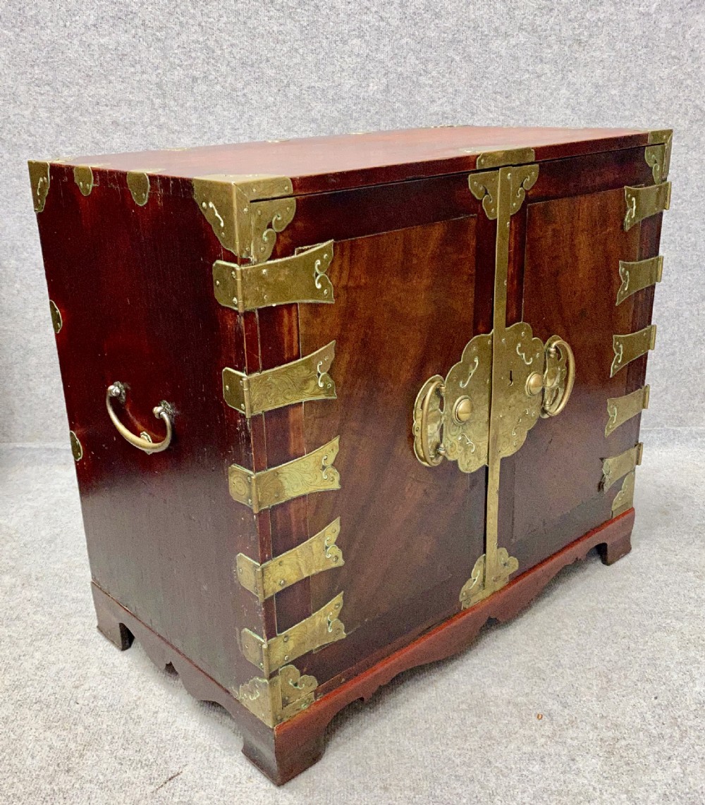 A small Chinese hardwood cabinet featuring polished dark wood, twin front doors, and decorative engraved brass mounts, hinges, and a central circular lockplate. View VIII