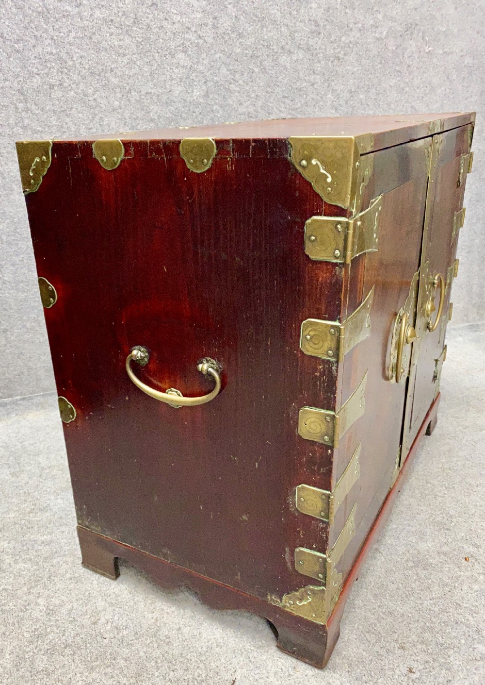 A small Chinese hardwood cabinet featuring polished dark wood, twin front doors, and decorative engraved brass mounts, hinges, and a central circular lockplate. View IX