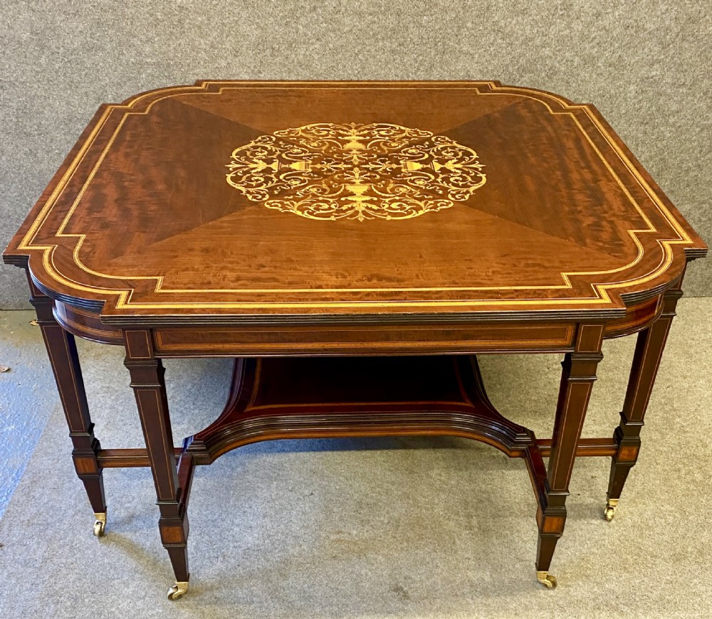 A square mahogany centre table featuring a central marquetry motif, indented corners, tapered legs, undertier, and brass castors.