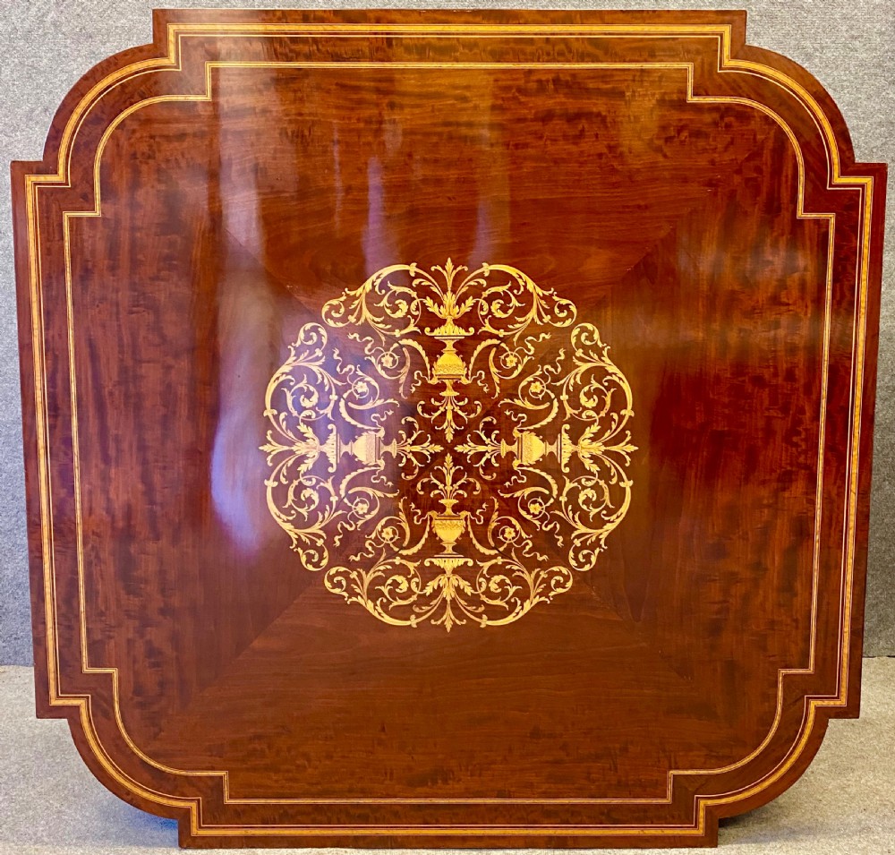 A square mahogany centre table featuring a central marquetry motif, indented corners, tapered legs, undertier, and brass castors. View II