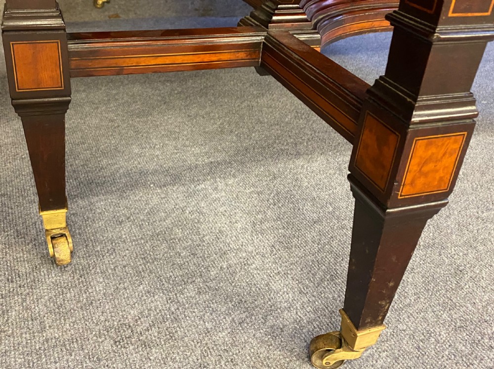 A square mahogany centre table featuring a central marquetry motif, indented corners, tapered legs, undertier, and brass castors. View VI