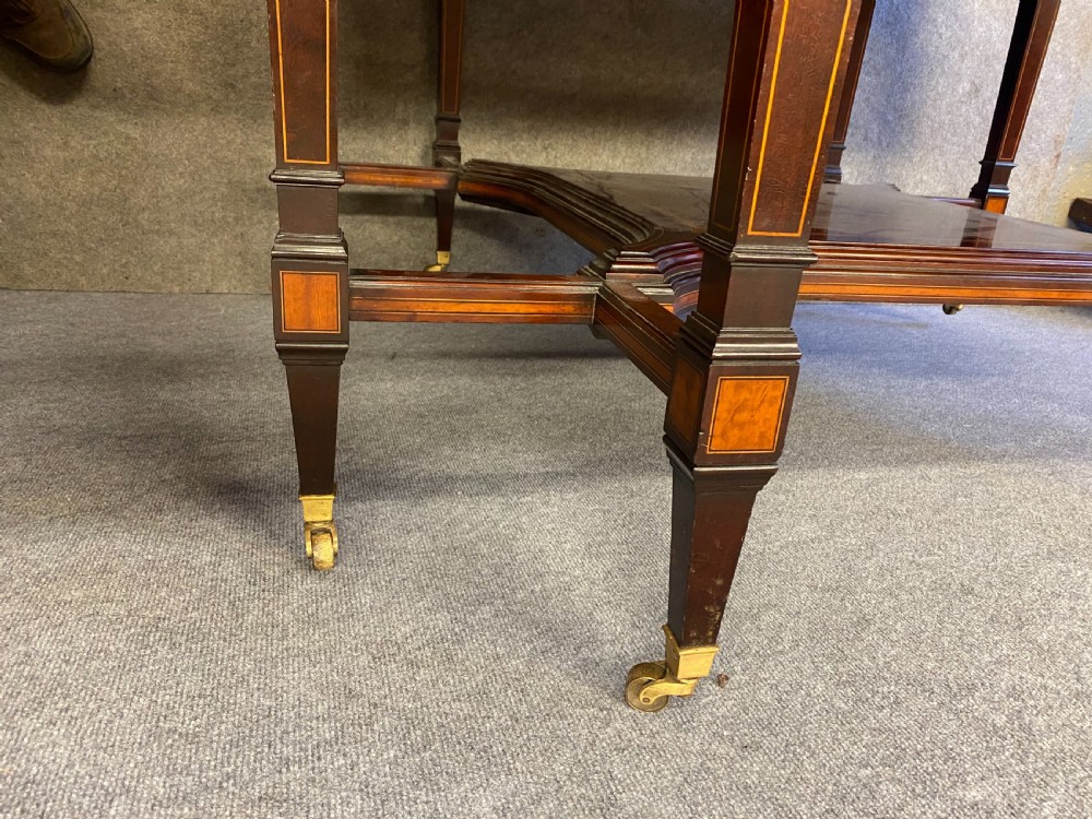 A square mahogany centre table featuring a central marquetry motif, indented corners, tapered legs, undertier, and brass castors. View VII