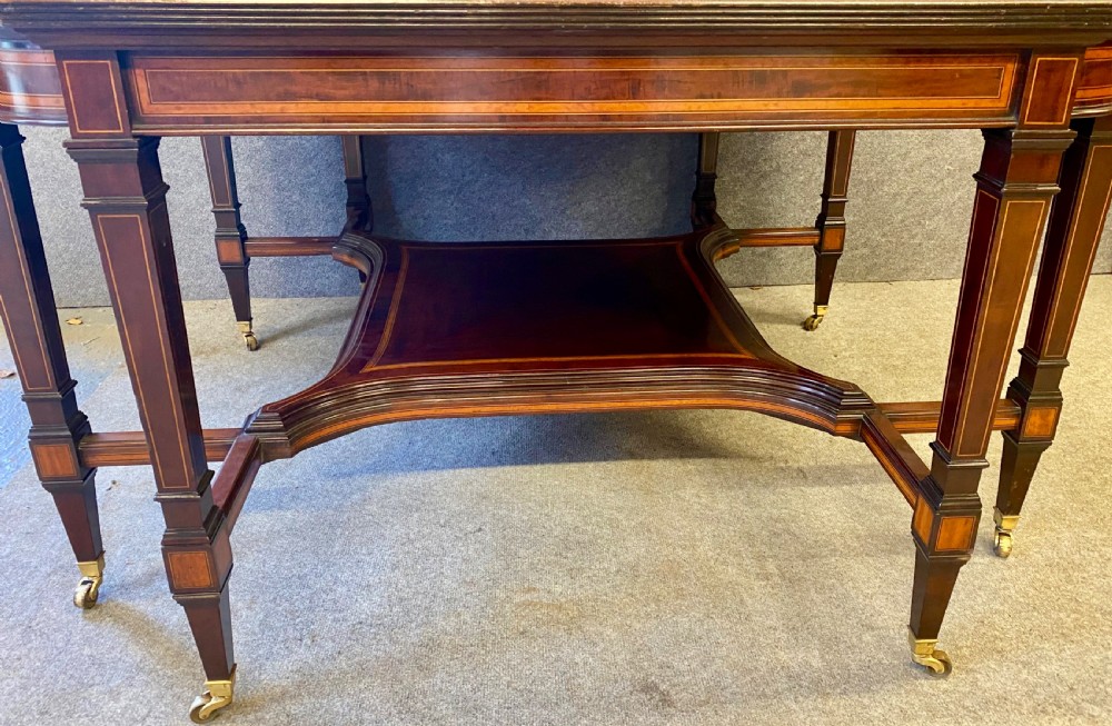 A square mahogany centre table featuring a central marquetry motif, indented corners, tapered legs, undertier, and brass castors. View VIII