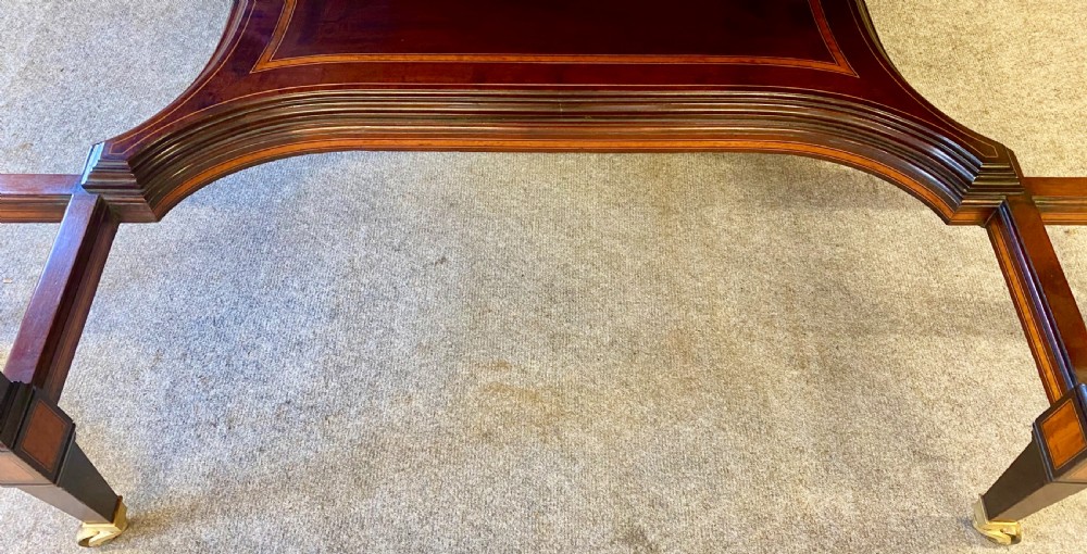 A square mahogany centre table featuring a central marquetry motif, indented corners, tapered legs, undertier, and brass castors. View IX