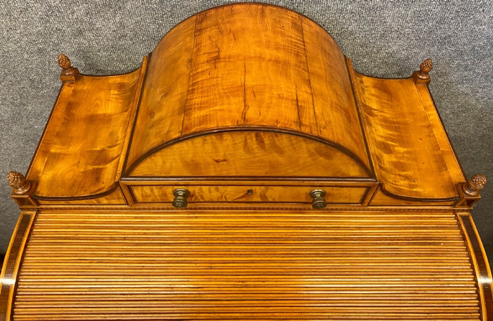 A George III satinwood cylinder desk featuring an arched cresting, a tambour roll top, two frieze drawers, and lower cupboard doors with oval inlay panels. View IV