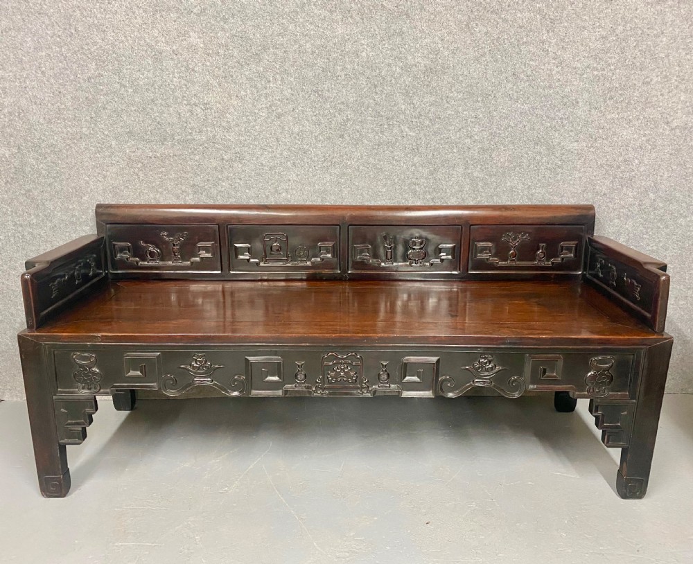 A dark hardwood Chinese bench featuring relief carved back panels depicting scholars objects, low side armrests, and an intricate apron with key fret motifs.