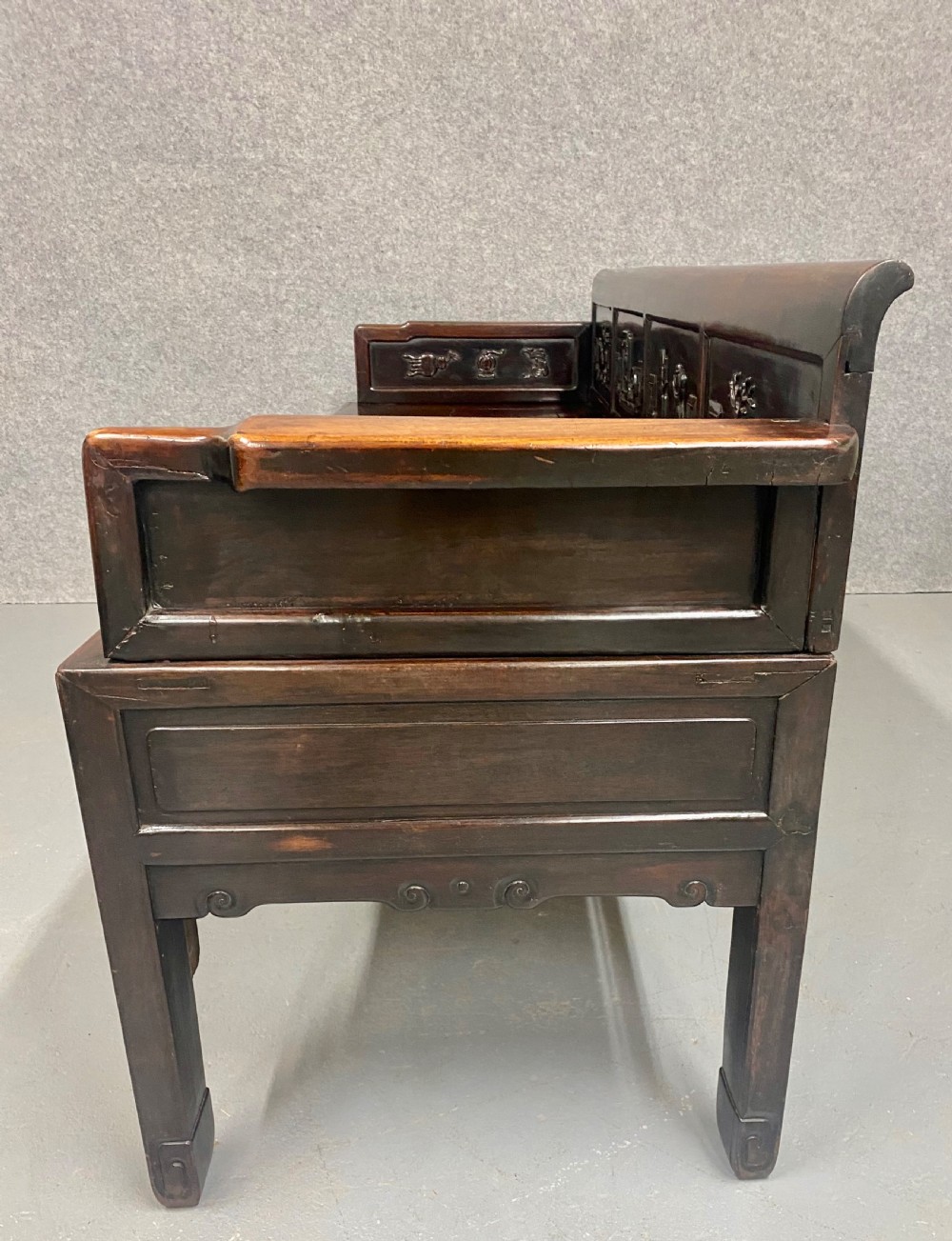 A dark hardwood Chinese bench featuring relief carved back panels depicting scholars objects, low side armrests, and an intricate apron with key fret motifs. View II