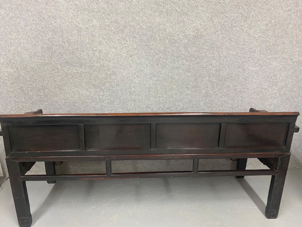 A dark hardwood Chinese bench featuring relief carved back panels depicting scholars objects, low side armrests, and an intricate apron with key fret motifs. View III
