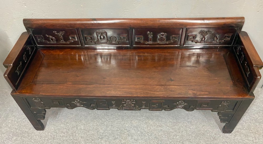 A dark hardwood Chinese bench featuring relief carved back panels depicting scholars objects, low side armrests, and an intricate apron with key fret motifs. View IV