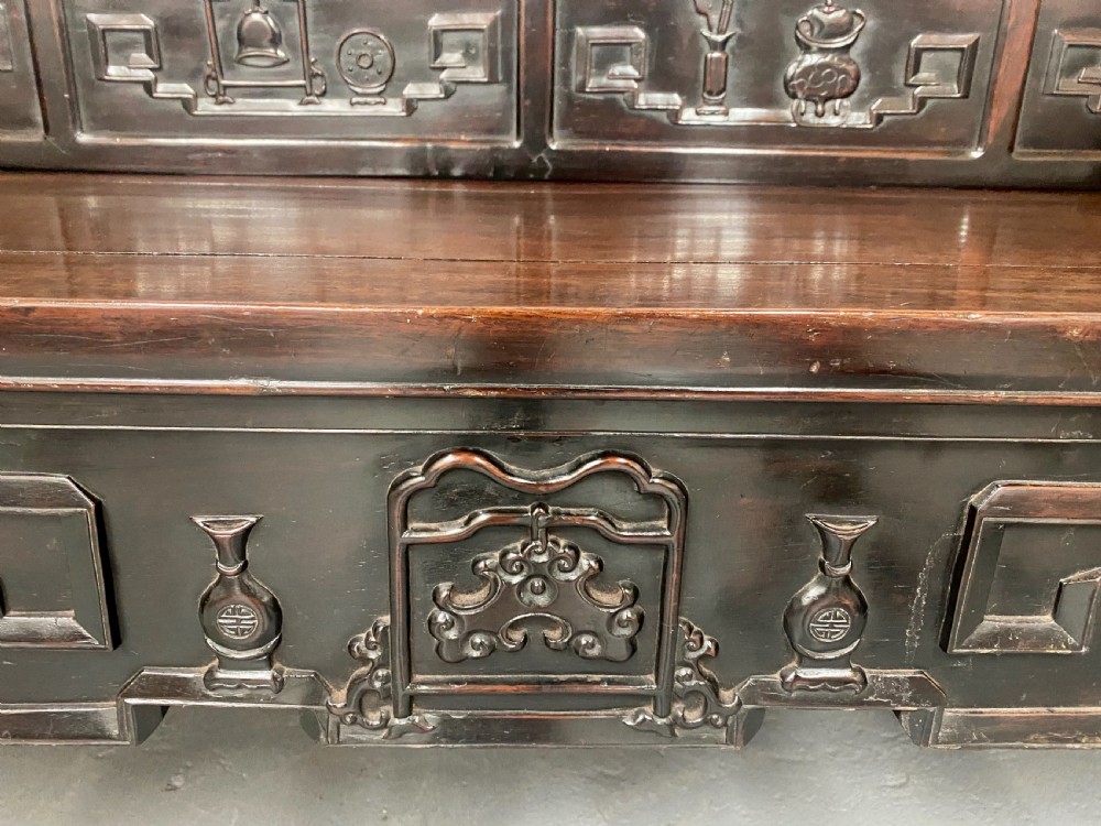 A dark hardwood Chinese bench featuring relief carved back panels depicting scholars objects, low side armrests, and an intricate apron with key fret motifs. View XII