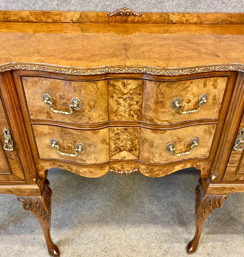 A nine-piece Queen Anne style dining room suite featuring a burr walnut dining table, matching sideboards, and red patterned fabric chairs with cabriole legs. View X