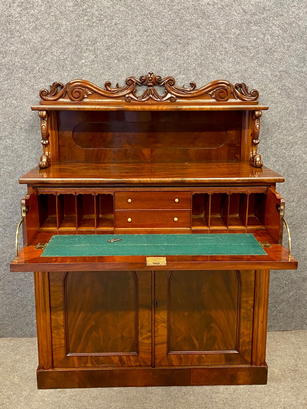 A Victorian flame mahogany secretaire chiffonier featuring an ornate carved cresting top shelf, drop-front desk compartment with green writing surface, and a two-door cupboard base.