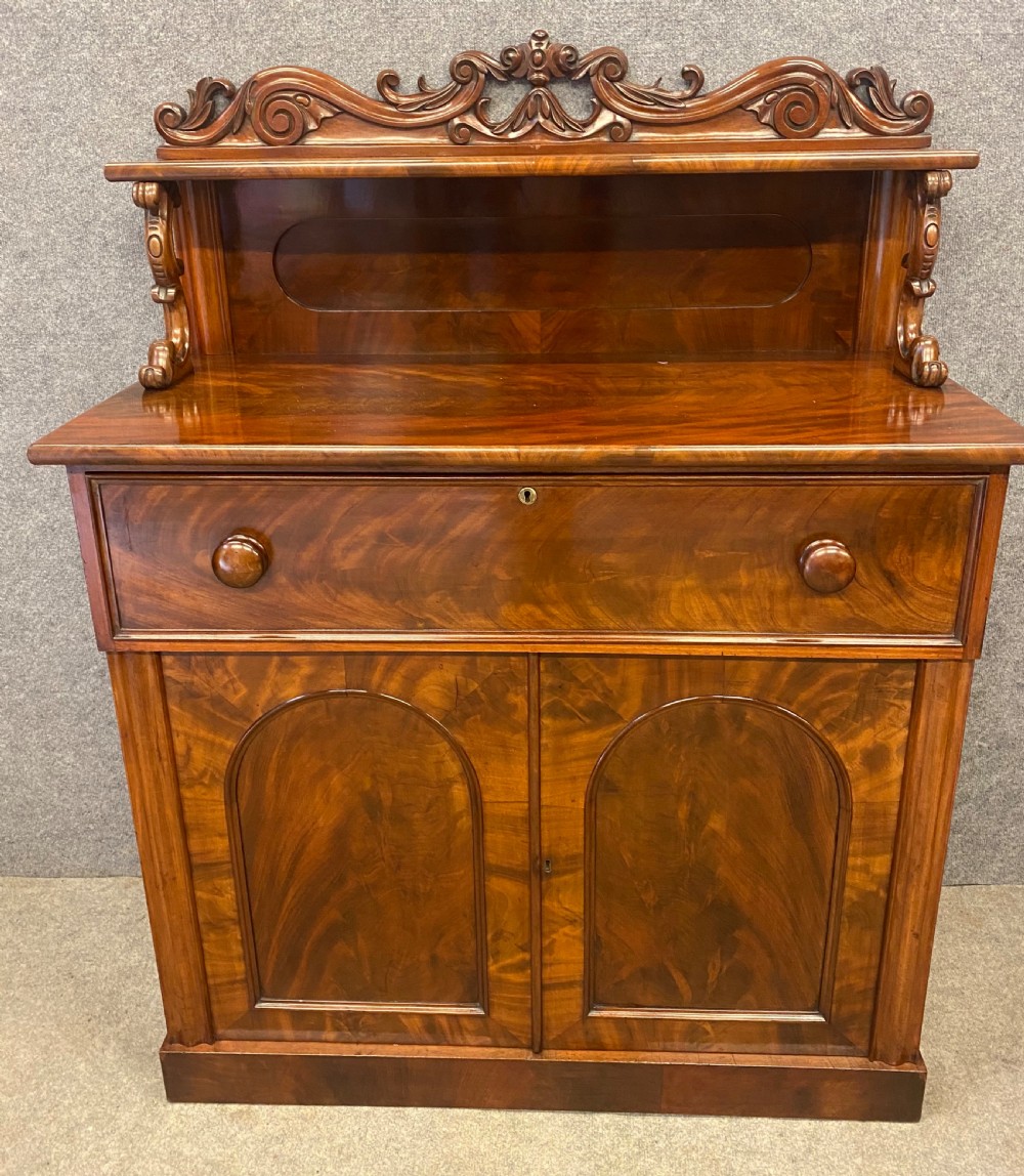 A Victorian flame mahogany secretaire chiffonier featuring an ornate carved cresting top shelf, drop-front desk compartment with green writing surface, and a two-door cupboard base. View II
