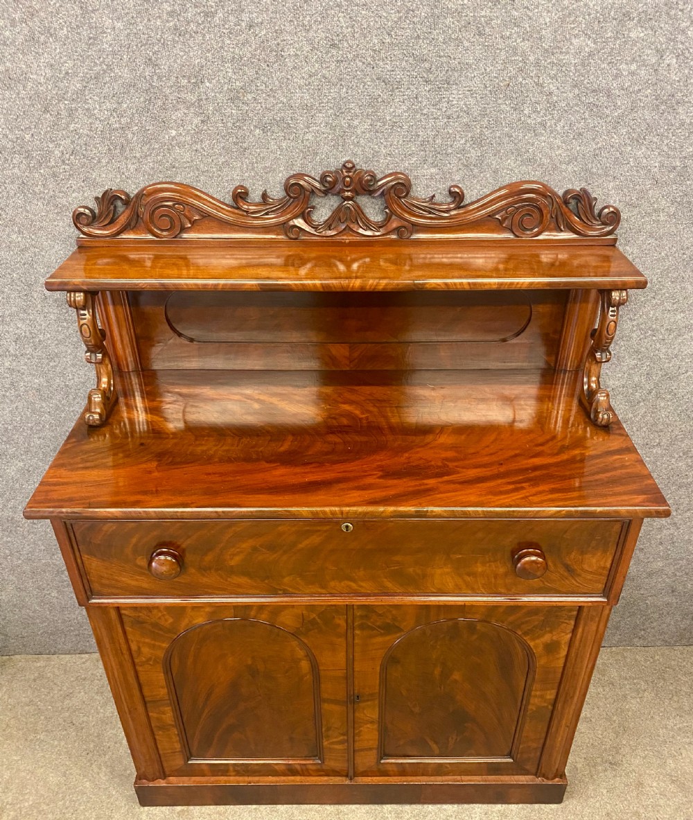 A Victorian flame mahogany secretaire chiffonier featuring an ornate carved cresting top shelf, drop-front desk compartment with green writing surface, and a two-door cupboard base. View III