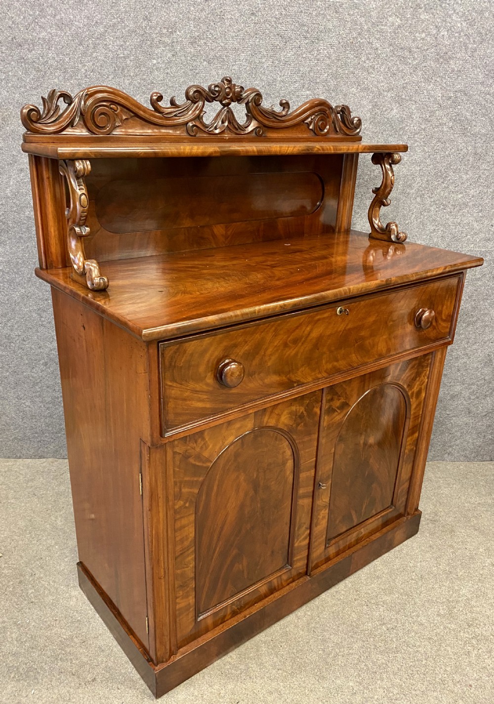 A Victorian flame mahogany secretaire chiffonier featuring an ornate carved cresting top shelf, drop-front desk compartment with green writing surface, and a two-door cupboard base. View IV